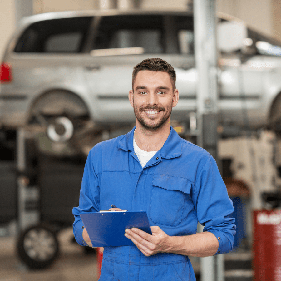 Smiling technician in blue overalls holding clipboard in automotive workshop with car on lift, representing reliable vehicle servicing and repairs.